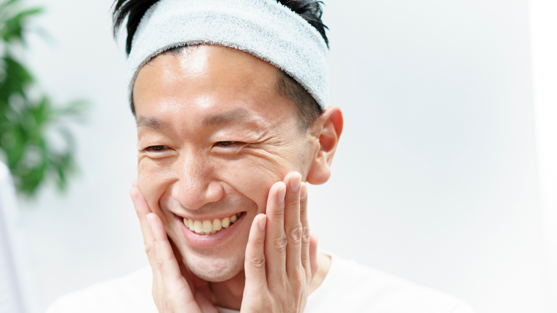 Smiling man with a headband massaging skincare product into his cheeks.
