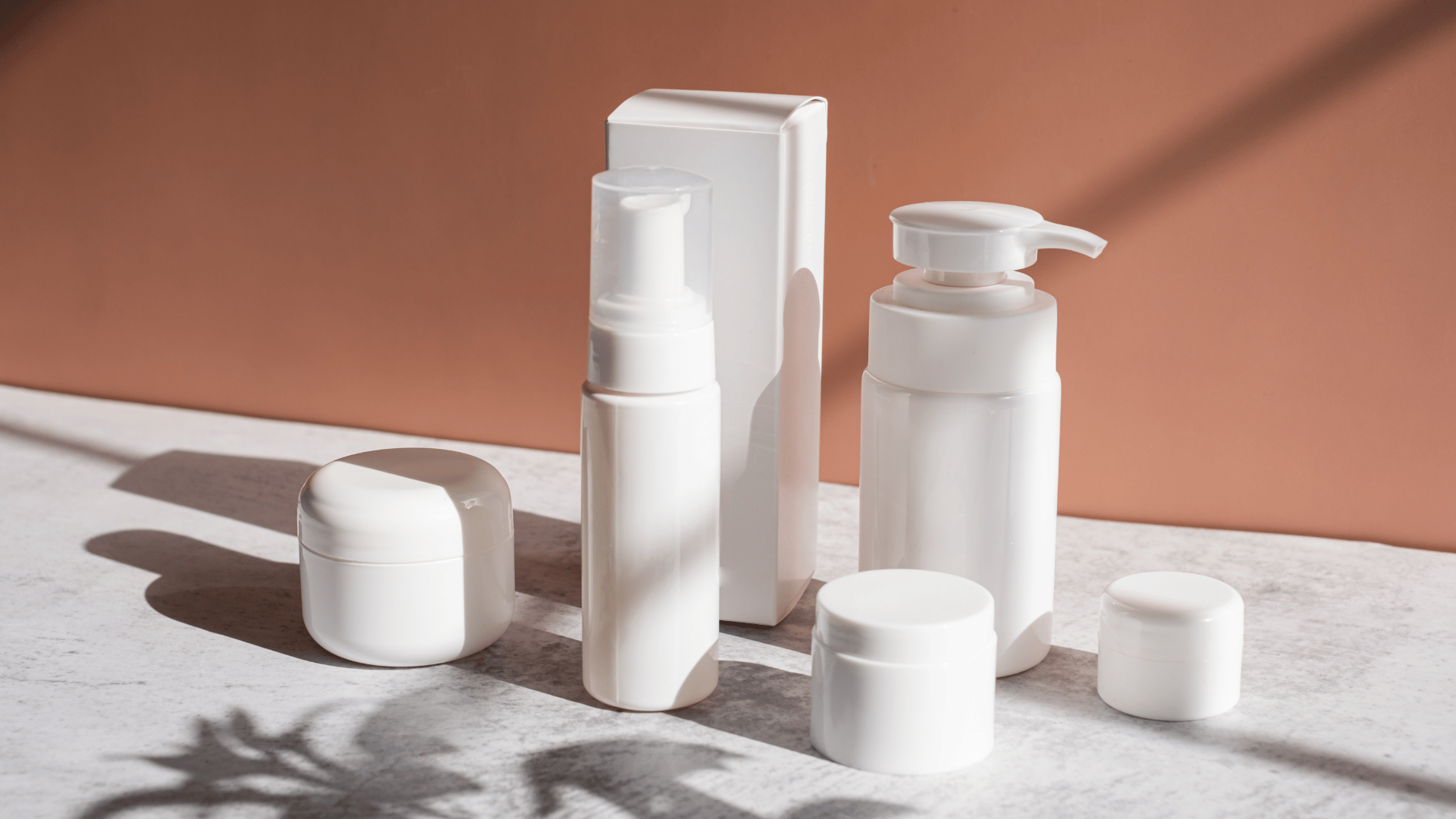 Minimal white skincare bottles and jars displayed on a marble surface in soft sunlight.