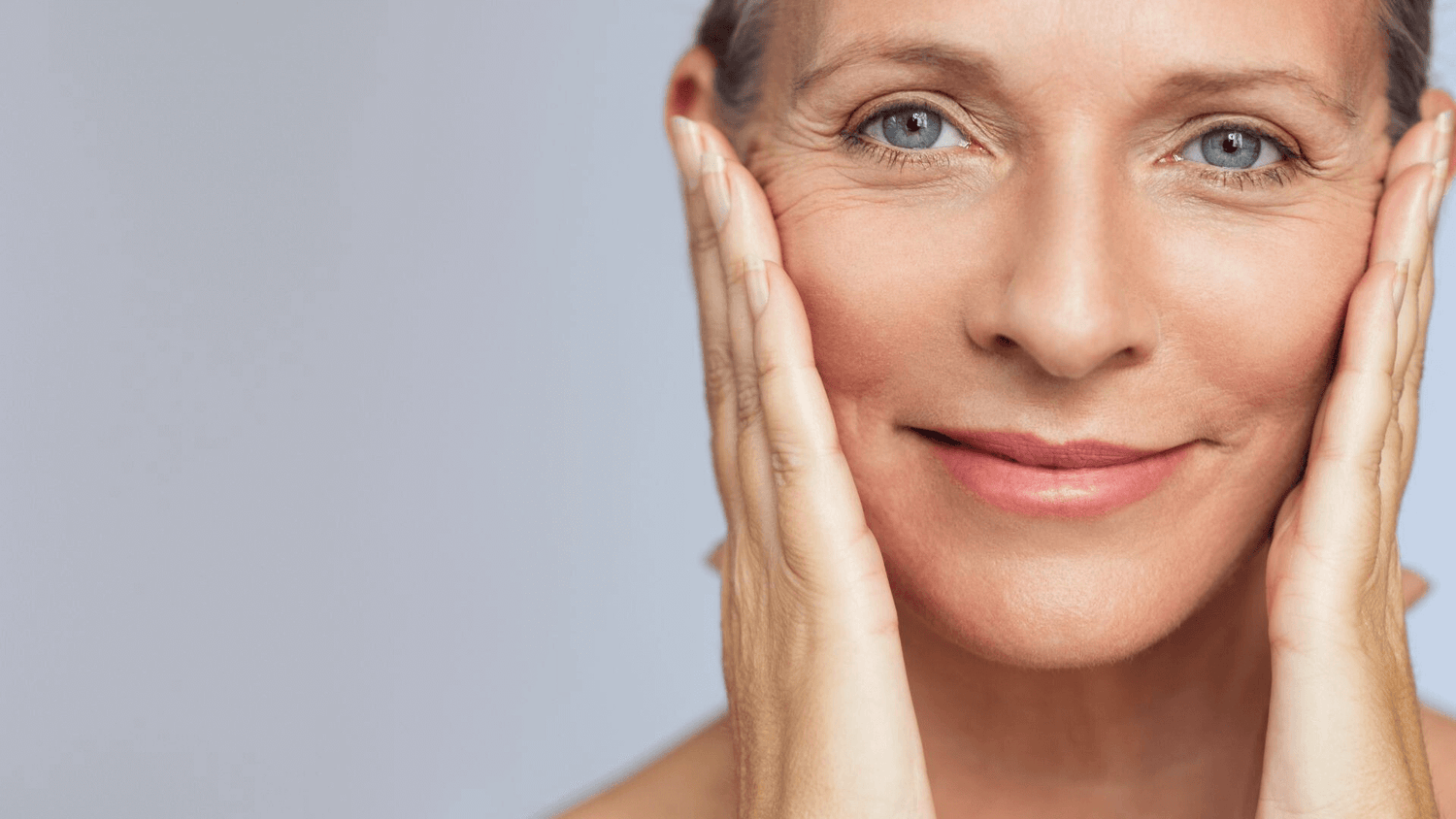 Mature woman gently holding her face with both hands and smiling at the camera.