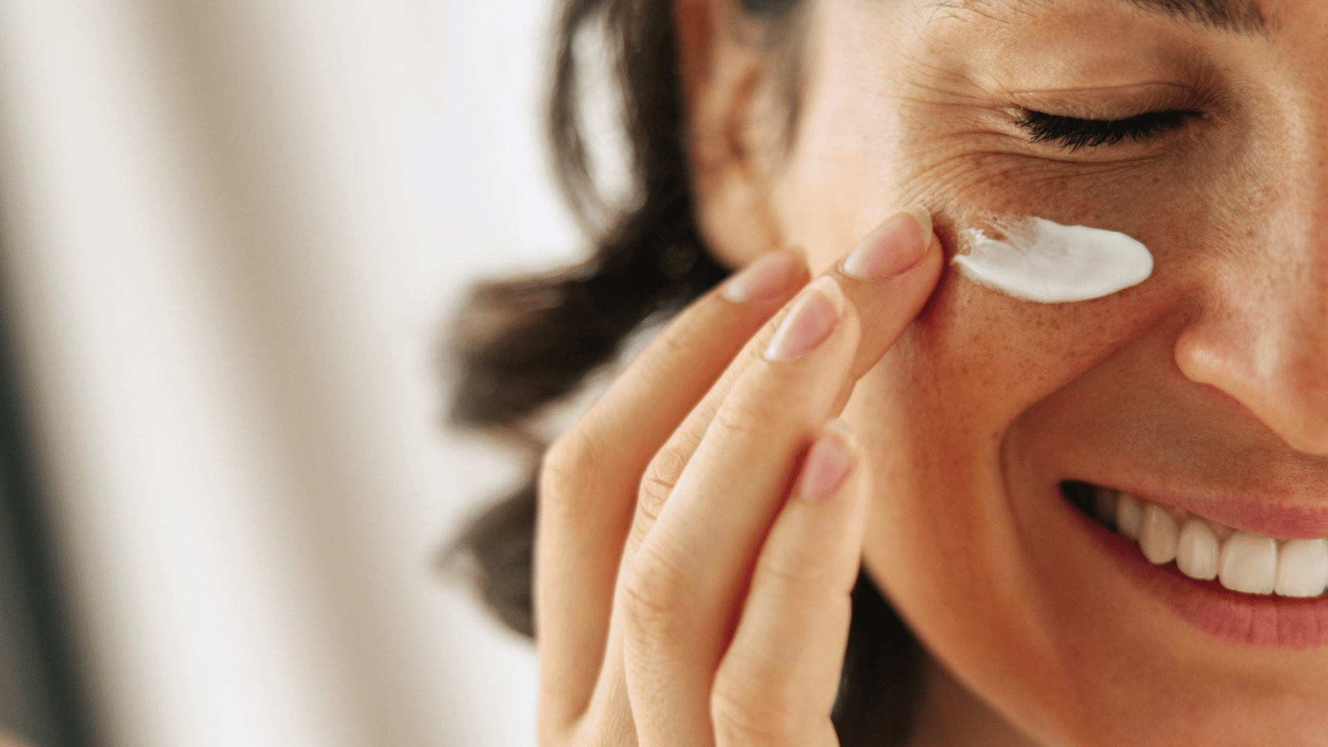 Smiling woman applying skincare cream under her eye with her fingertip.