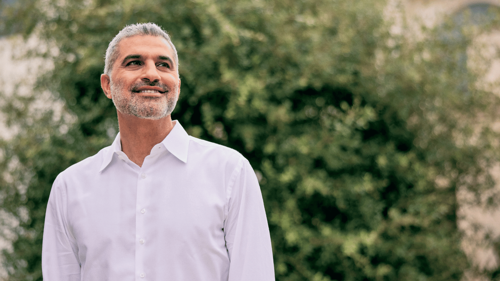 Dr. Karam creator of KaramMD Trifecta standing in a white shirt in front of a green foliage background, healthy and smiling