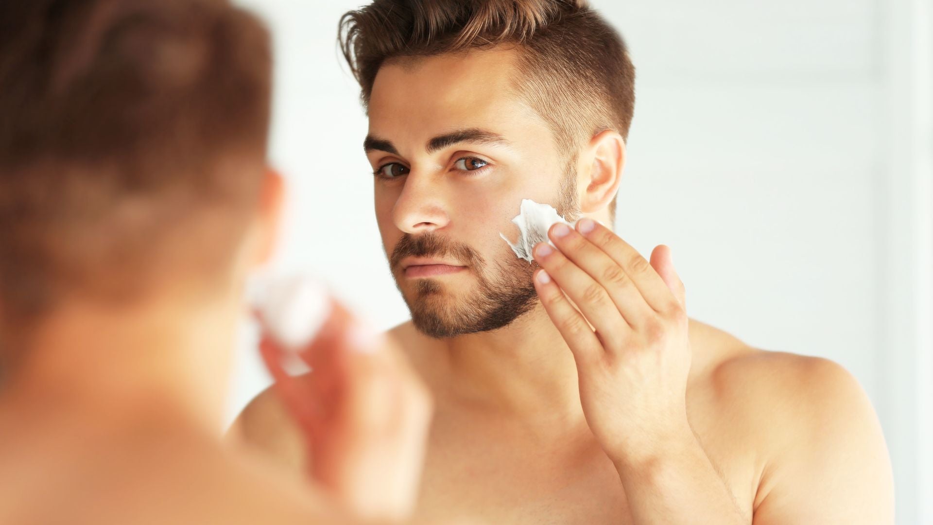 Man applying skincare cream to his cheek while looking in the bathroom mirror.